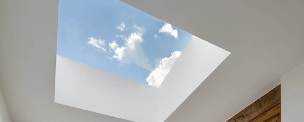 A cozy interior space featuring a skylight that illuminates the room. Large windows showcase a green landscape and cloudy sky. A patterned rug covers the floor, and plush seating is arranged along the wall.