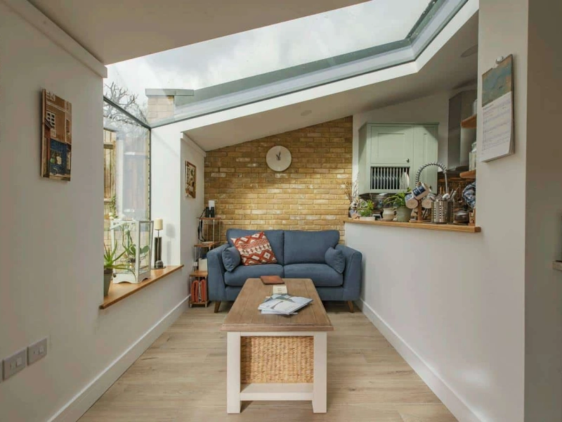 A kitchen with a blue sofa with a clock above it. To the left there is a eaves skylight which is letting natural light in
