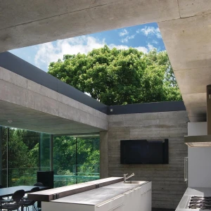 A modern kitchen features a sleek white island with drawers and a stainless steel sink, surrounded by large glass windows that overlook lush greenery. The ceiling is made of concrete, allowing natural light to flood the space. A wall-mounted television is visible in the background.