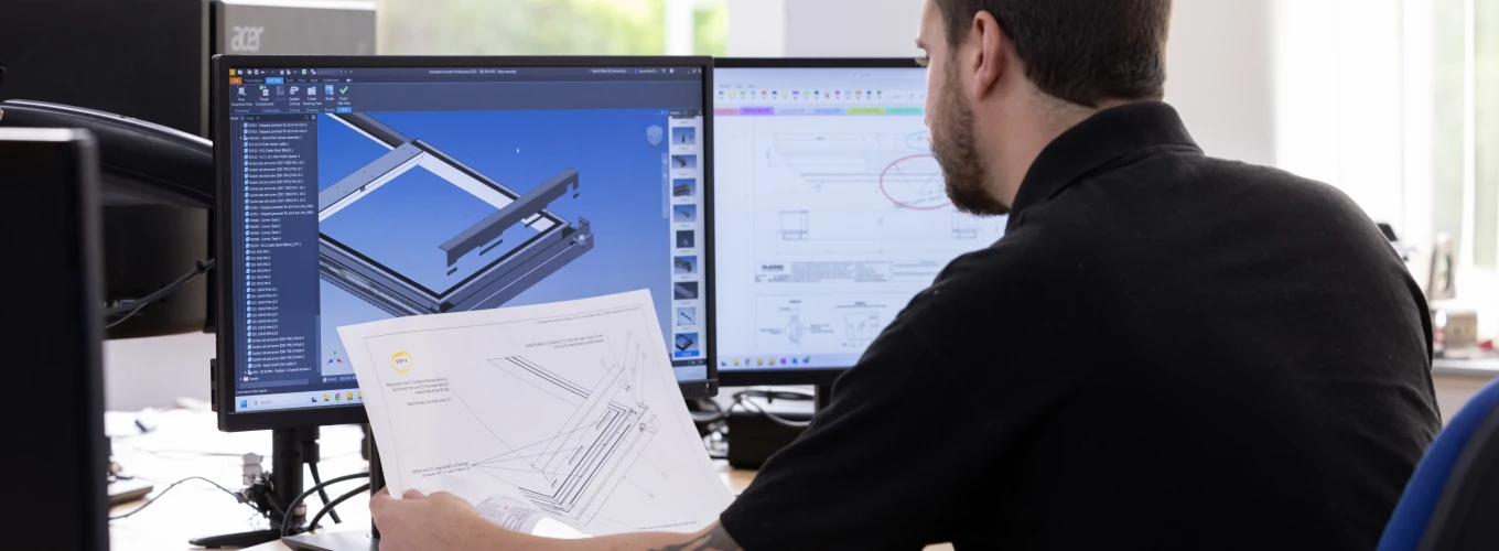 A man sits at a desk, examining a blueprint while looking at two computer monitors displaying technical designs. Natural light streams through a large window in the background.