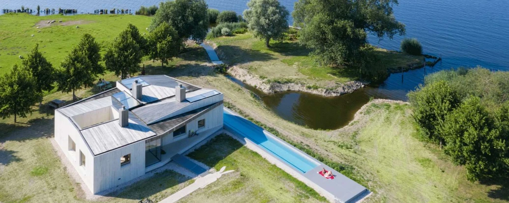 A modern house with a flat roof sits near a large body of water, surrounded by green grass and trees. A long, narrow swimming pool extends beside the house, leading to a serene landscape with calm water and distant wind turbines.