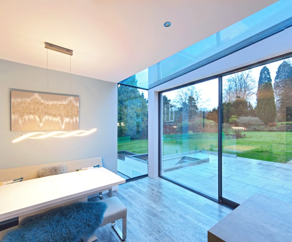 A modern dining area featuring a sleek white table and soft seating, with a large glass wall providing a view of a lush garden outside. Soft lighting hangs above the table, creating a warm atmosphere.