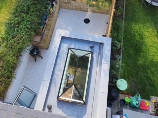 Aerial view of a landscaped backyard featuring a stone patio, a circular drain, a wooden fence, and lush greenery. There's a glass skylight visible on the roof and a trampoline in the corner.