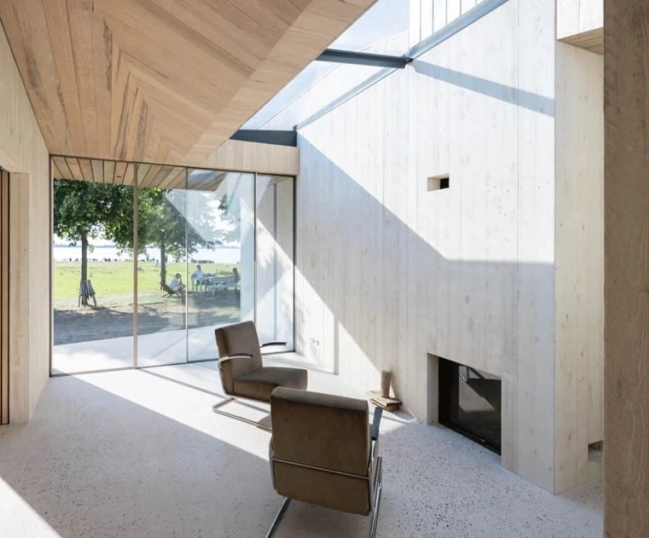 A modern interior featuring light-colored wooden walls and a sloped ceiling. Large glass windows provide a view of an outdoor space with greenery. A minimalist chair and a fireplace are positioned within the room, creating a serene atmosphere.