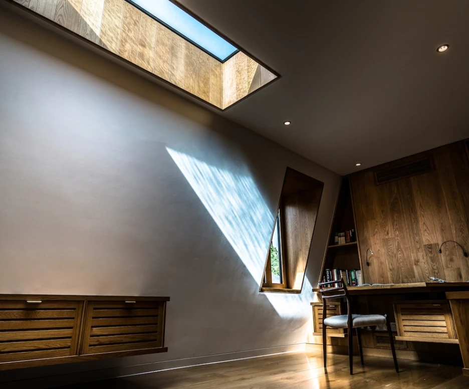 A modern interior featuring a wooden desk and a chair beside a wall with a triangular window. Light from a skylight casts a geometric shadow on the wall, creating a warm atmosphere. Shelves filled with books are visible in the background.