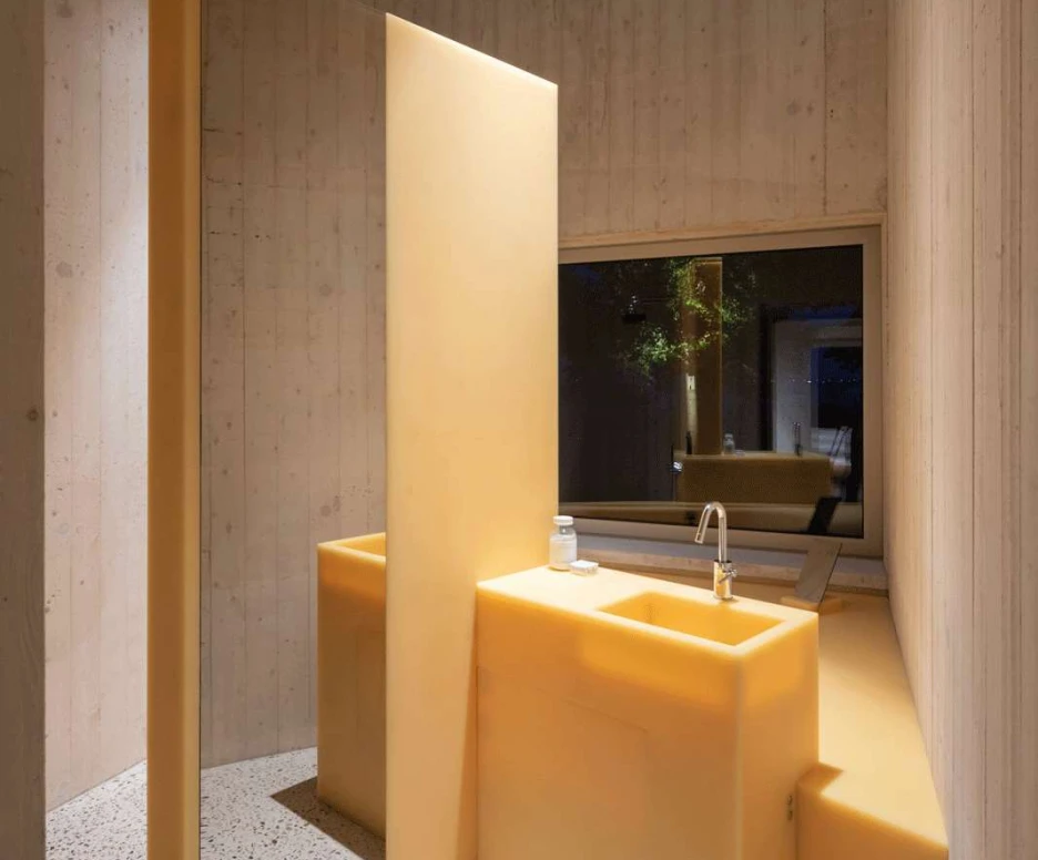 A modern bathroom featuring bright yellow walls and sink area, with a large mirror and a window allowing natural light. The floor has a textured finish, and the ceiling is painted blue, creating a vibrant atmosphere.