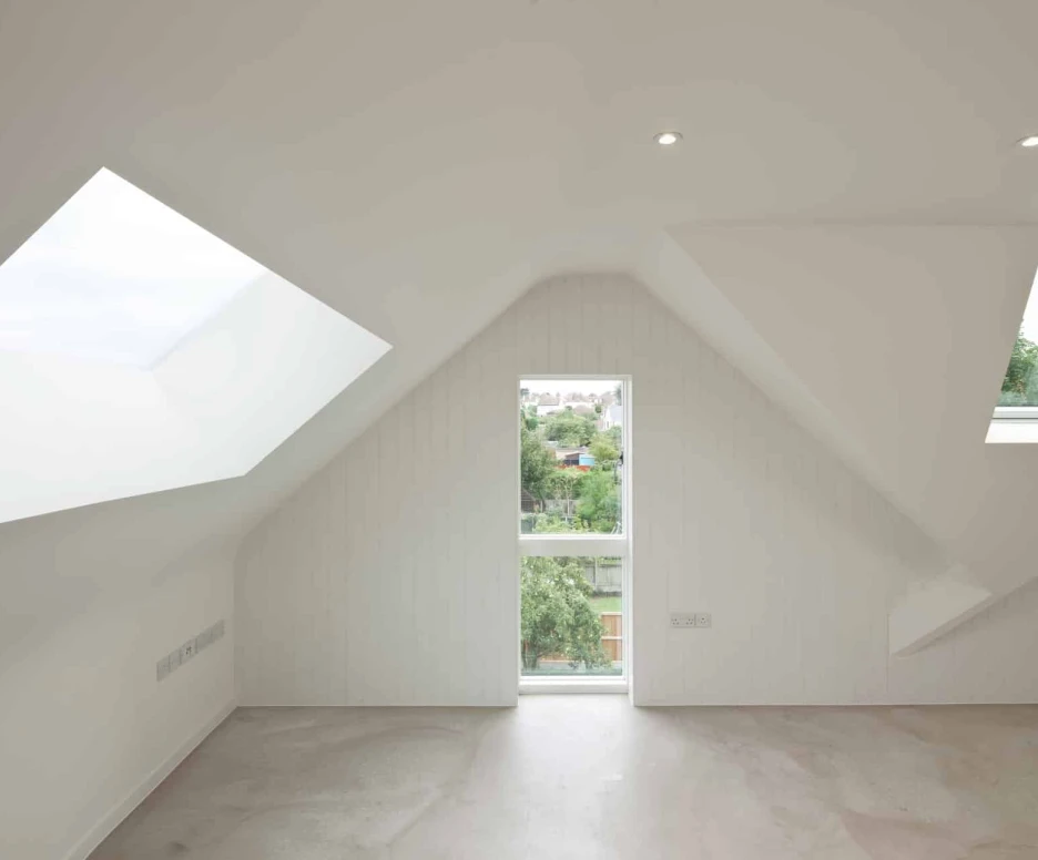 A modern, minimalist room with sloped white walls and angular windows. Natural light filters in from a large skylight and a vertical window, revealing greenery outside. The floor features a light-colored, polished surface.