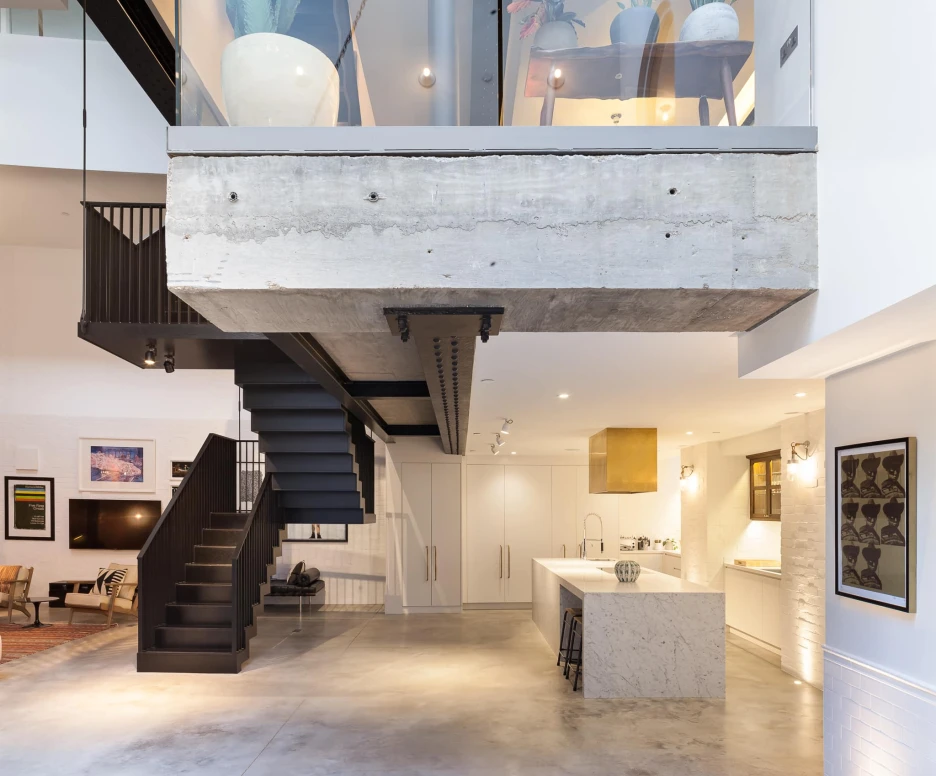 A modern interior space featuring a concrete architectural feature, a sleek staircase, and a kitchen with a marble island. The room has high ceilings with large windows and plants, creating a bright and airy atmosphere. Stylish furniture is arranged for comfort.