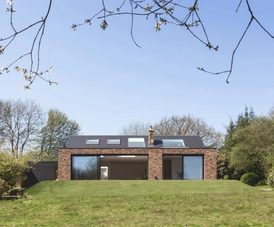 A modern, low-profile brick house is set against a clear blue sky, with large windows showcasing an open interior. Surrounding the house are lush green lawns and trees in the background.