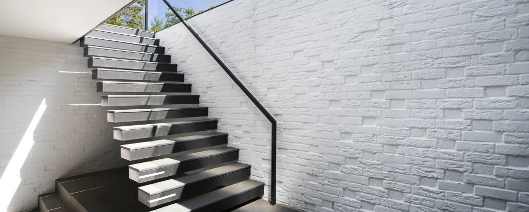 Modern staircase leading upwards, featuring sleek wooden steps and a glass railing. The walls are textured and white, with natural light coming in from above, illuminating the space.
