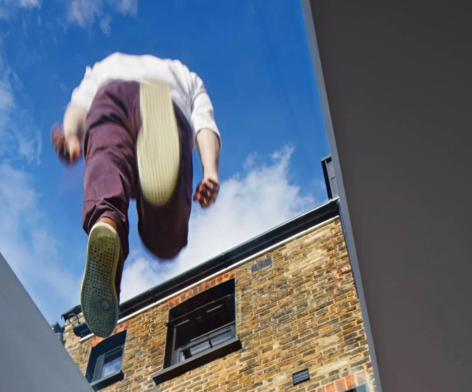 A person is jumping through a skylight, seen from below, with legs extended downward and a clear blue sky in the background. A brick building is visible behind them.