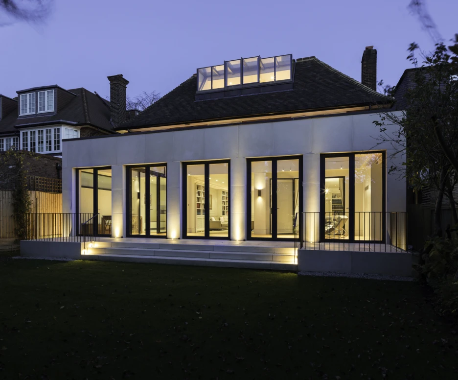 A modern house at dusk, featuring large glass windows that illuminate the interior. The structure is surrounded by a green lawn and has a wooden fence in the background. The roof has a skylight, adding to its contemporary design.
