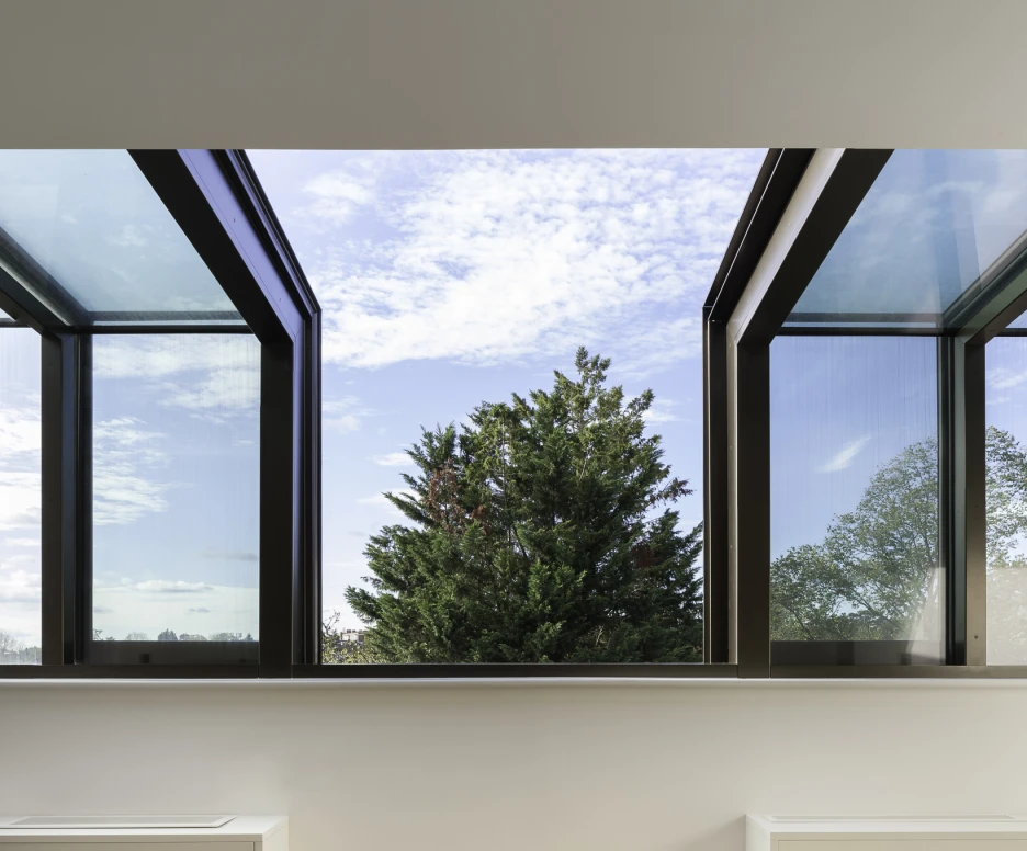 Large windows with a modern design open up to a view of a green tree and blue sky with scattered clouds. The interior features light-colored walls, wooden flooring, and minimalist furniture.