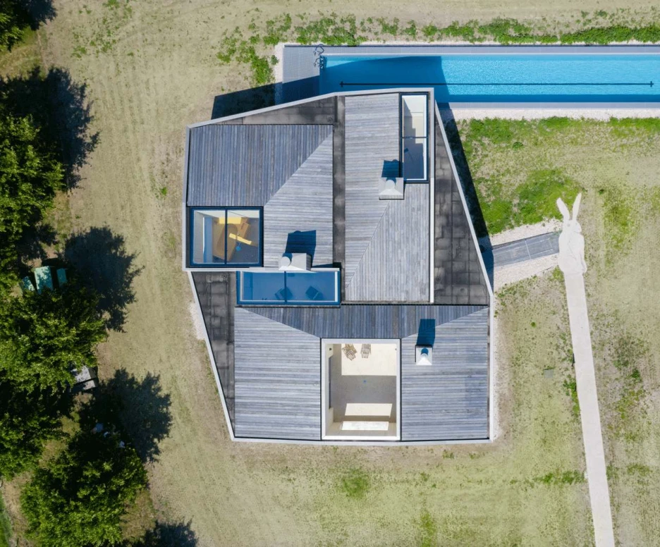 Aerial view of a modern, angular house featuring a flat roof and wooden deck. A rectangular swimming pool runs alongside the house, surrounded by a grassy lawn.