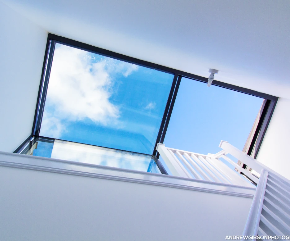 A view looking up through a large skylight, revealing a bright blue sky with a few fluffy clouds. The interior features white walls and a staircase leading to the skylight.