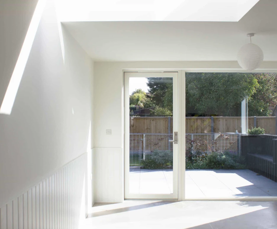 Bright, airy interior space featuring a sliding glass door that opens to a balcony. Natural light streams in from a skylight, illuminating the minimalist white walls. A lush garden can be seen outside, framed by the door.