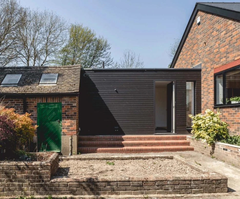 A modern, dark wooden extension attached to a brick house, featuring steps leading to an open door. In the foreground, a landscaped area with a low brick wall and minimal vegetation, along with a green shed on the left. Trees and blue sky are visible in the background.