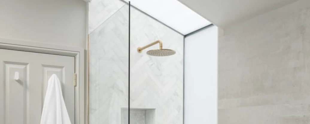 A modern bathroom featuring a glass shower enclosure with a gold showerhead and fixtures. The walls are tiled with light-colored marble in a herringbone pattern. A white towel hangs on the door. Natural light enters through a skylight above the shower.