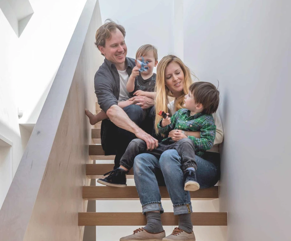 A family of four sits on a staircase. A father and mother hold their young children, enjoying a playful moment. The interior features modern, minimalist design with natural light coming from a skylight above.
