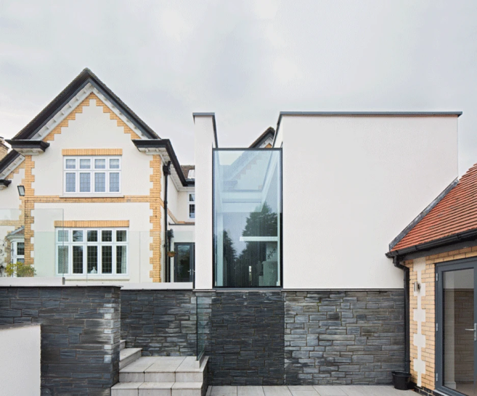 A modern building with a glass extension is situated alongside a traditional brick house. The structure features a mix of slate and smooth white surfaces, with stone steps leading up to the entrance.