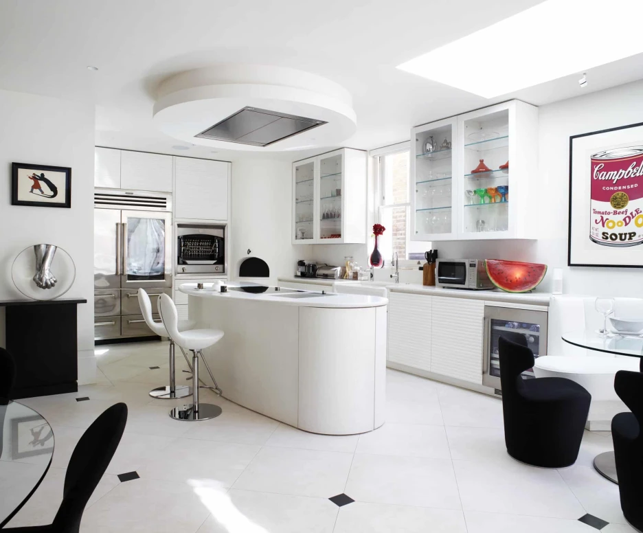 A modern kitchen featuring sleek white cabinetry, a large island with bar stools, and a stylish dining area. Bright natural light streams in through large windows, highlighting decorative glassware and a Campbell's Soup poster on the wall.