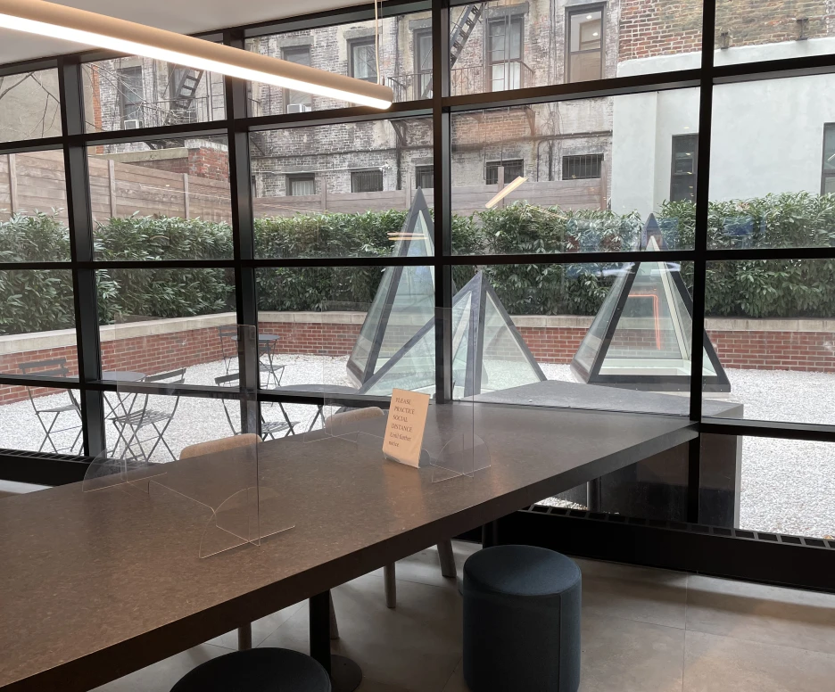 A modern indoor space features a large table with protective dividers, overlooking a courtyard with geometric glass installations and outdoor seating. Sunlight streams through large windows, creating a bright atmosphere.