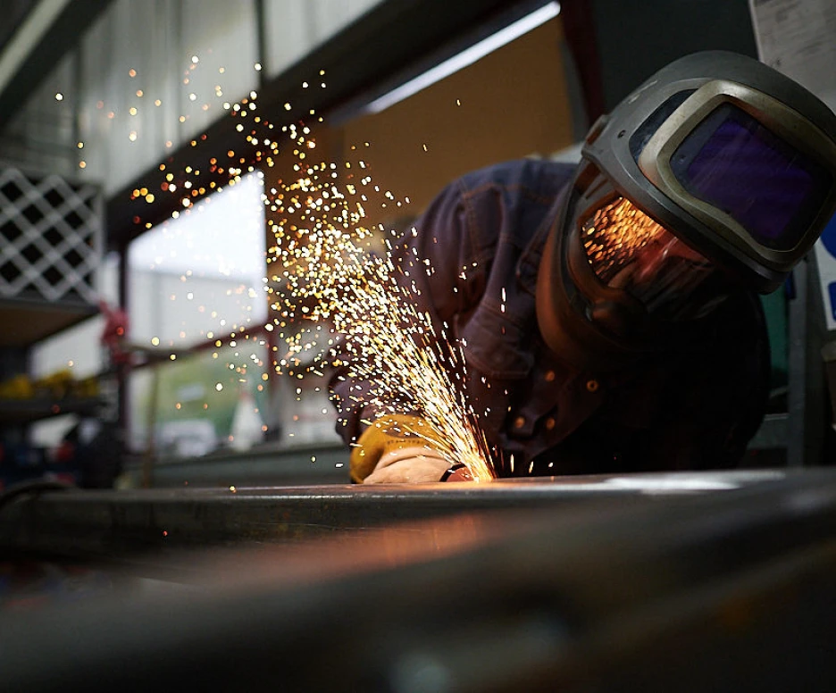 A person wearing a welding helmet and protective gear is working on metal, creating sparks as they weld. The background features industrial equipment and tools.