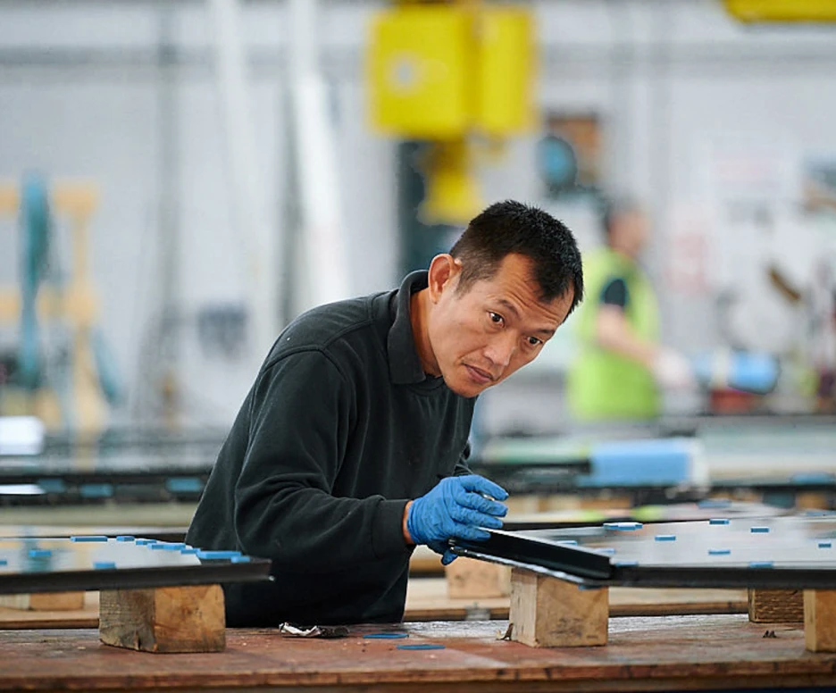 A man wearing a dark sweater and blue gloves works intently at a manufacturing facility, handling large sheets of material on a wooden table. The background features equipment and other workers engaged in similar tasks.