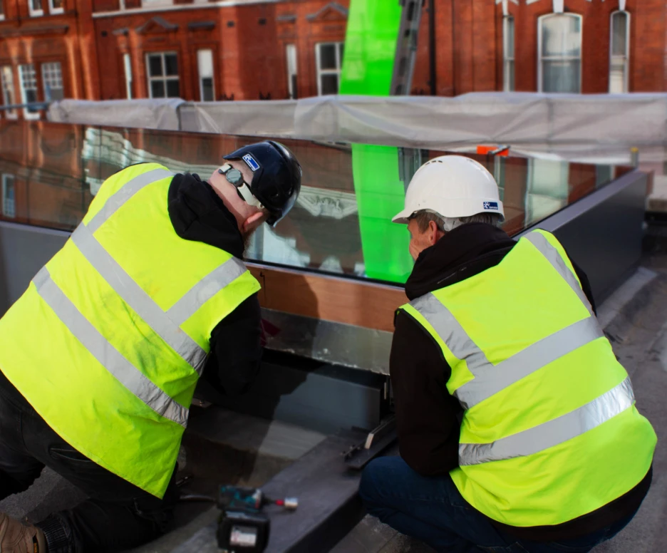 Two construction workers, wearing safety vests and helmets, are kneeling and working on a reflective metal surface. They are on a rooftop, with a green tarp visible in the background and red brick buildings. Tools are scattered around them.