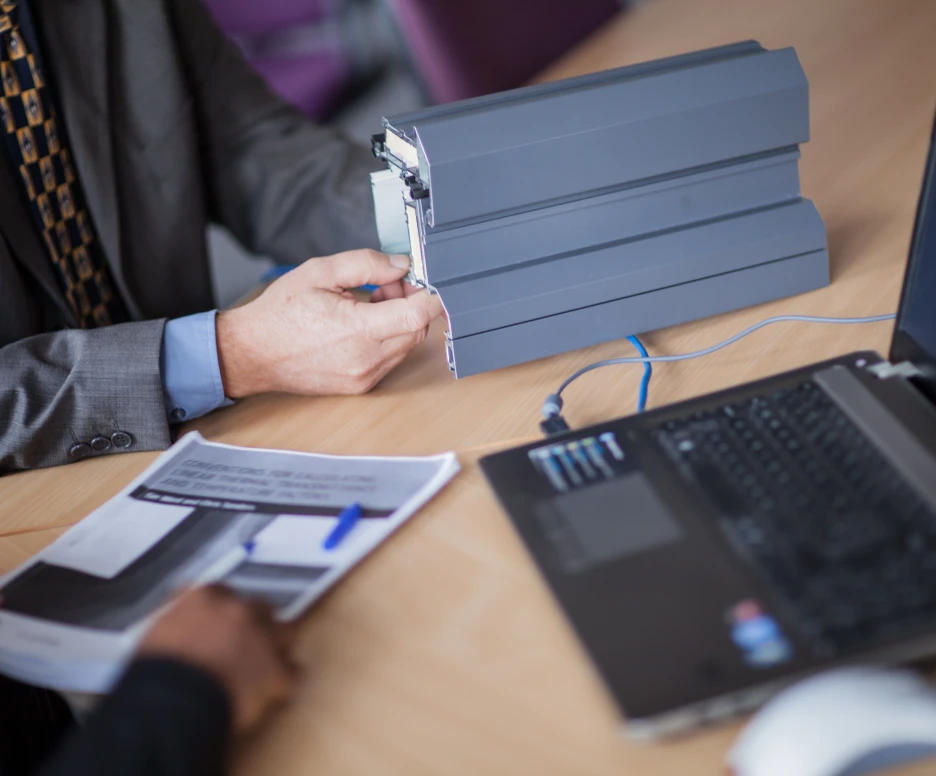 A business professional with a gray suit examines a gray device, while another person holds printed documents beside a laptop on a conference table.