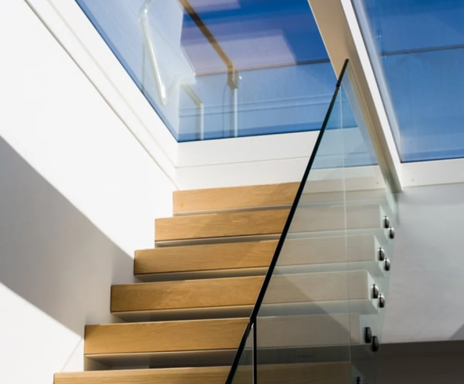 A modern staircase with wooden steps and a glass railing, leading to a bright space with a skylight. Sunlight casts shadows on the wall and steps.