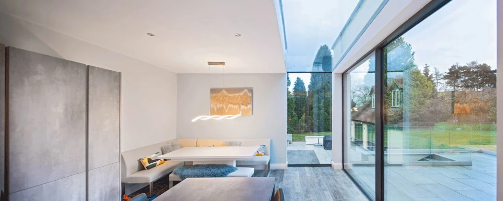 A modern dining area with a minimalist design features a built-in seating nook, a sleek table, and large glass windows showcasing a green outdoor space. Neutral colors dominate the interior, and a stylish light fixture hangs above the table.