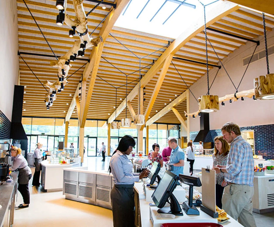 A modern café with a wooden ceiling and large windows. Several people are engaging at the counter, ordering food and drinks. Light fixtures hang from the ceiling, adding a warm ambiance. Various food items are displayed in the background.