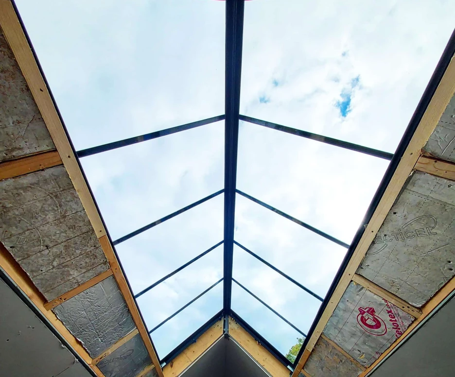 A skylight frames a view of the cloudy sky from inside a building under construction. The wooden frame is partially insulated, showcasing exposed materials and structure.