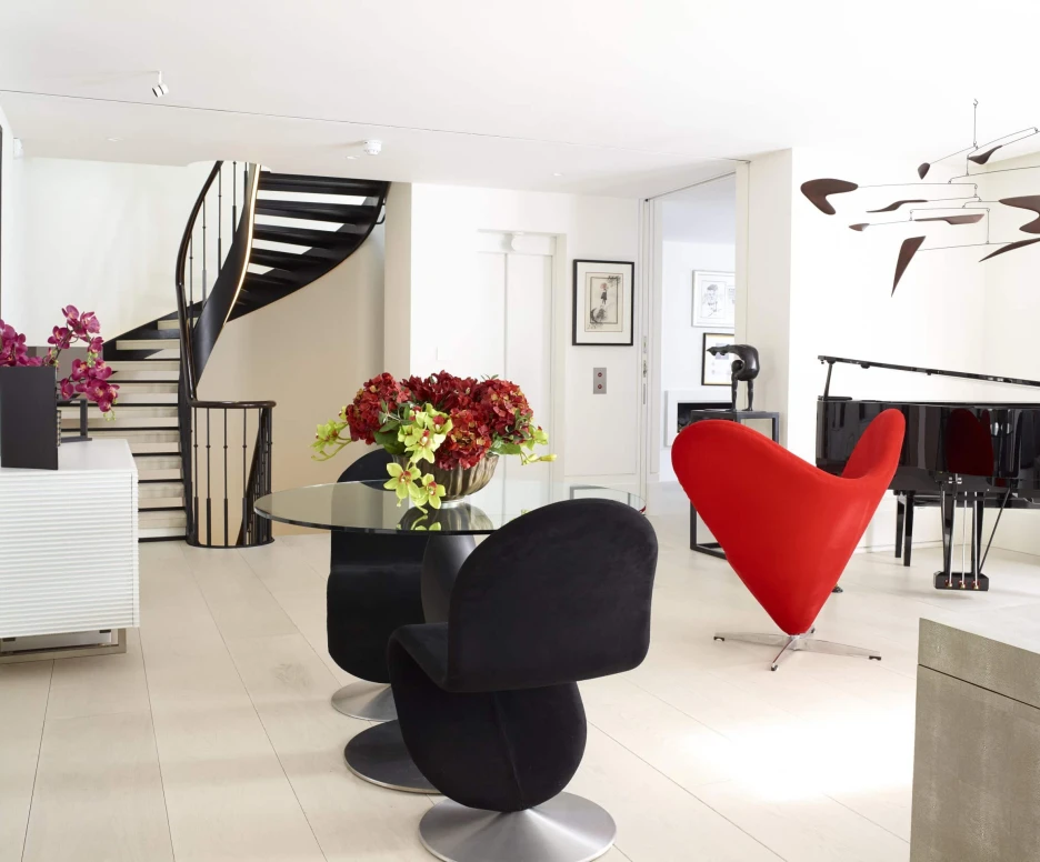 A modern living space with a distinctive heart-shaped red chair, a round glass dining table surrounded by black chairs, and vibrant flowers in vases. A spiral staircase and a grand piano are visible in the background, adding elegance to the bright, open layout.