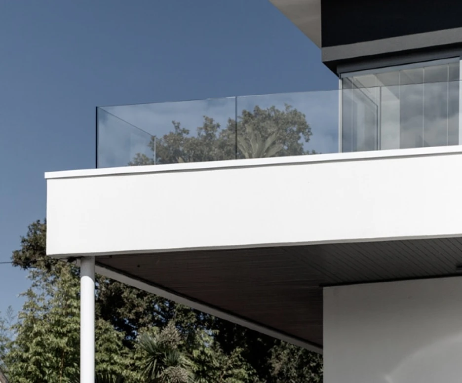 A modern house featuring a sleek balcony with glass railing, set against a clear blue sky and greenery in the background.