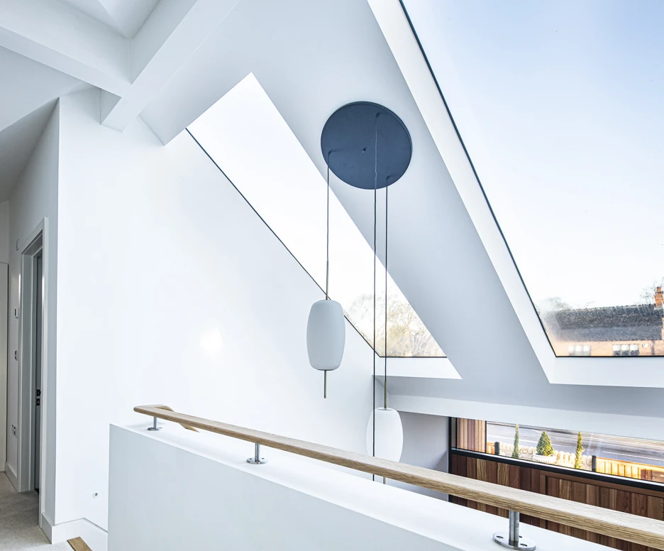 Bright, modern interior featuring a spacious hallway with a skylight and minimalist design. Light walls and a wooden railing complement the contemporary aesthetic. A circular pendant light hangs from the ceiling, illuminating the space.