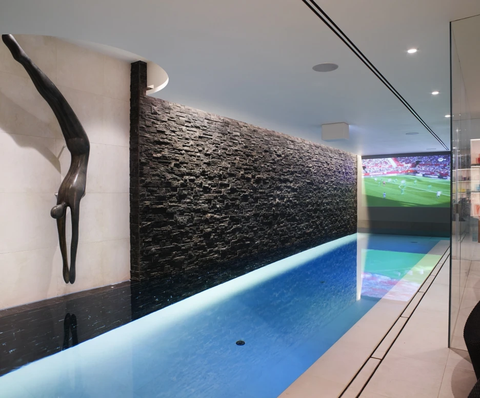 A sleek indoor swimming pool with a modern design. A stone wall features a sculpture of a diver, while a screen displays a sports game in the background. The pool has clear blue water and contemporary lighting.