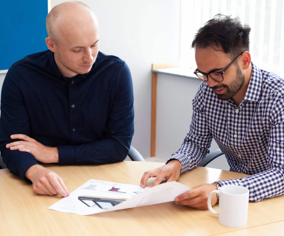 Two men are seated at a table, reviewing printed documents. One person is wearing a blue shirt and has short hair, while the other wears a checkered shirt and glasses. A white mug is on the table beside them.