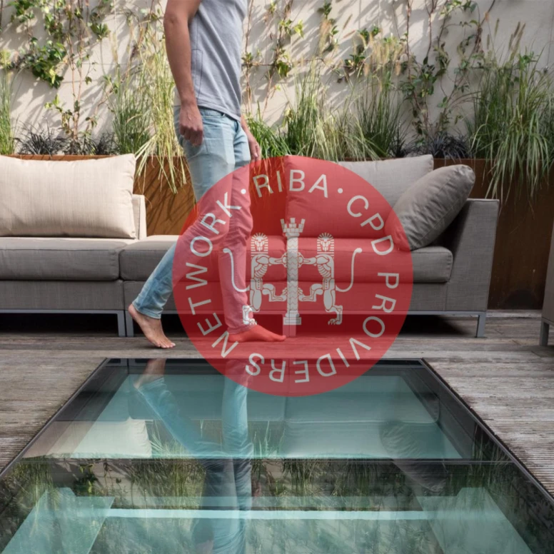 A person walking over a glass floor panel, revealing a lower level beneath, while surrounded by an outdoor seating area with plants and modern furniture.