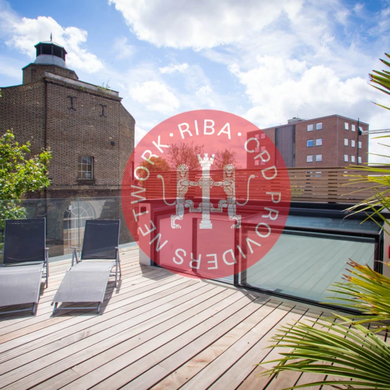 A rooftop terrace with wooden decking, featuring two lounge chairs and greenery. In the center, a red circle with a logo that includes the text "NETWORK. RIBA. CRD PROVIDERS." and two lion figures. Clouds are visible in the blue sky.