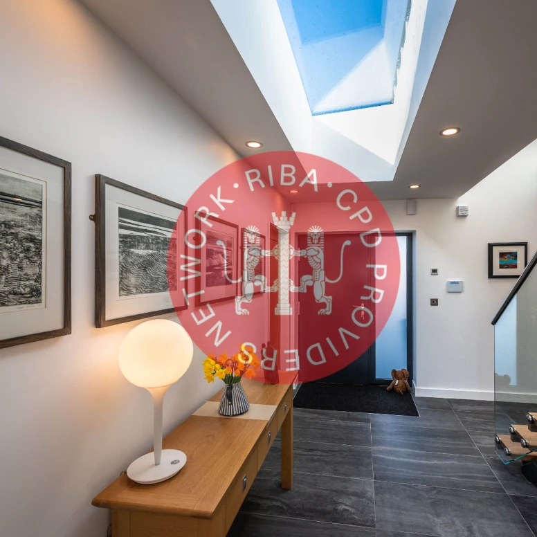 A modern hallway featuring a wooden console table with a round lamp, framed artwork on the walls, and a skylight flooding the space with natural light. A small table displays a vase of flowers, and a staircase is visible in the background.