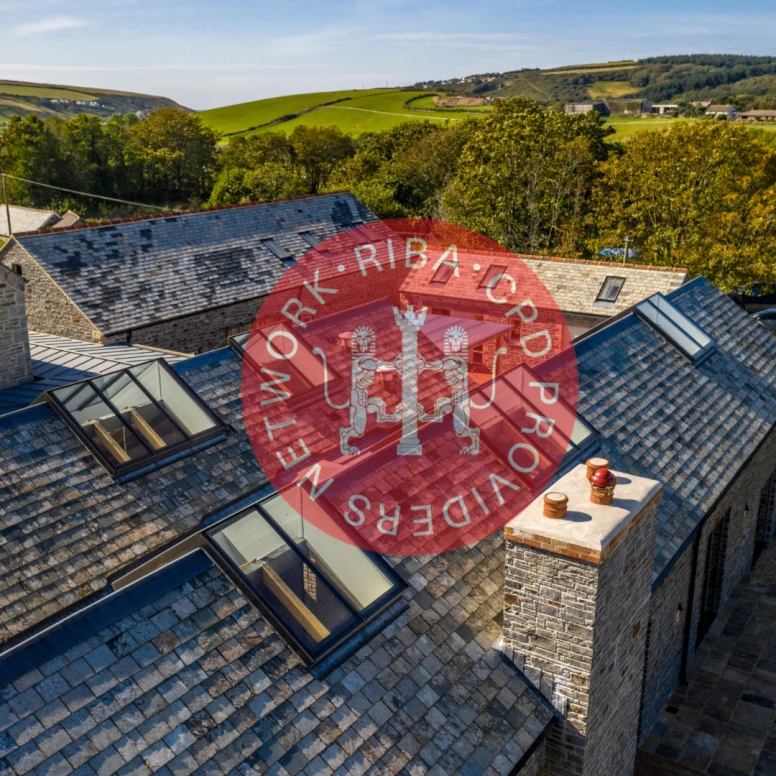 Aerial view of a stone building with a slate roof, surrounded by green hills and trees. There are several skylights on the roof and a chimney. A logo overlay is present in the center.