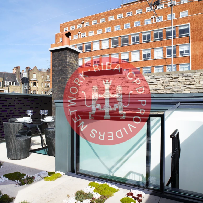 A modern rooftop terrace featuring a glass railing, a small dining area with two chairs and a table, and a landscaped garden area with moss and small plants. In the background, a red brick building is visible.
