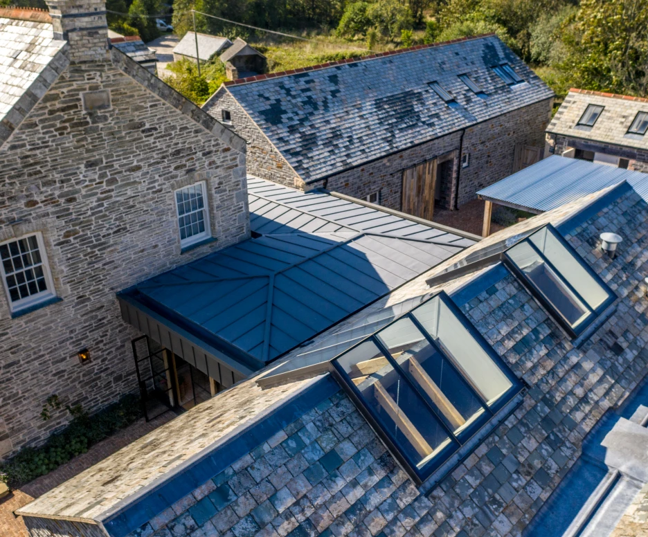 Aerial view of a rustic stone house and several nearby buildings, featuring a slate roof and modern skylights. Surrounding landscape includes trees and open space.