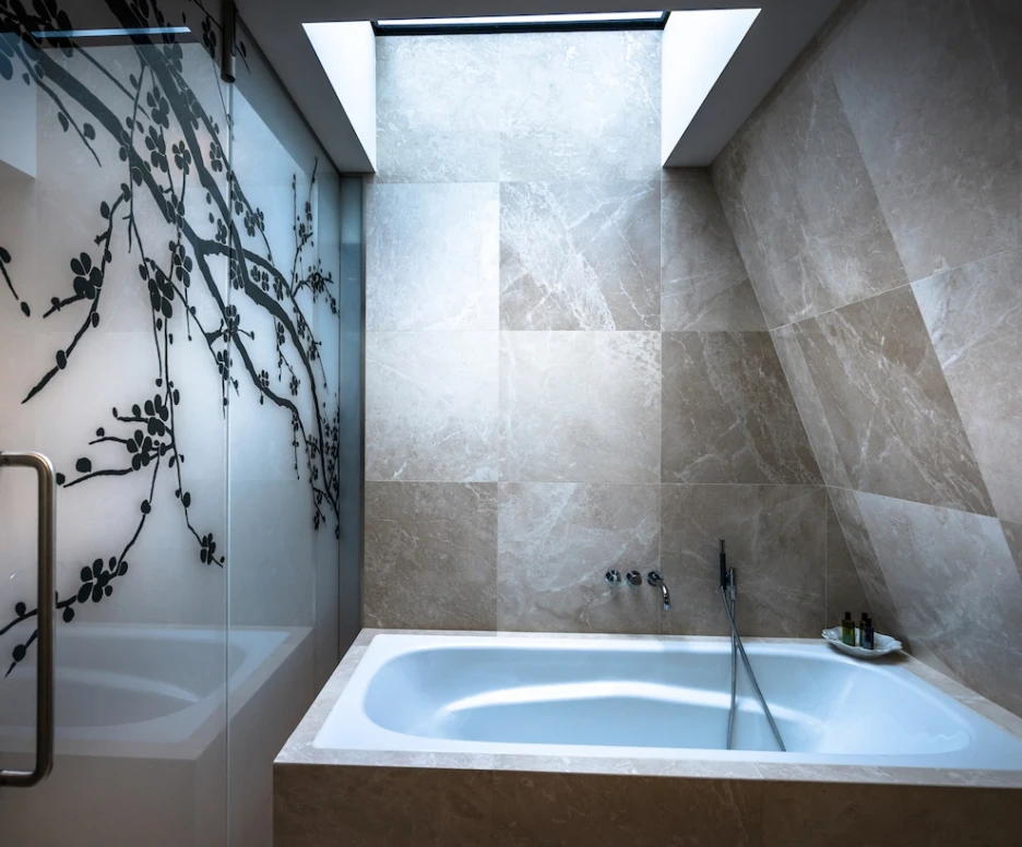 A modern bathroom featuring a sleek white bathtub against a backdrop of light-colored marble tiles. A glass shower with a decorative black leaf design is visible, and natural light pours in from a skylight.