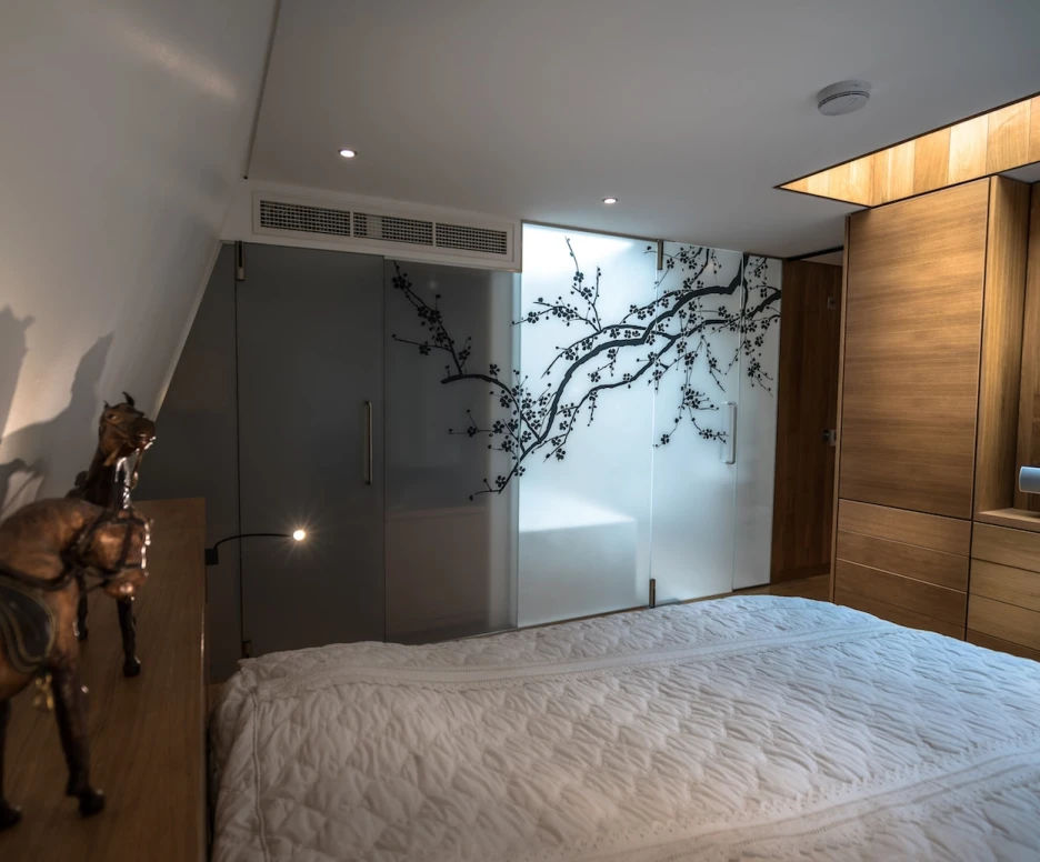 A stylish bedroom featuring a bed with a white quilt, wooden furniture, and modern decor. A glass wall adorned with a black floral design separates the sleeping area from a bathroom. Decorative figurines of horses sit on the bedside table.