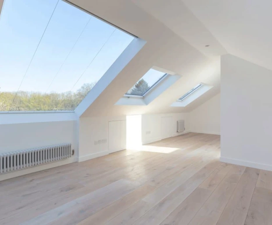 Bright, airy attic space with slanted skylights, white walls, and light wooden flooring. Minimalist design with a radiator along one wall.