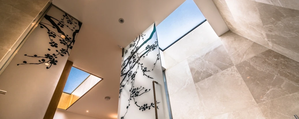 A modern bathroom with a sleek design featuring large stone tiles, a glass wall with artistic branch designs, and skylights allowing natural light to enter. The view is from below, highlighting the shower area and the visual harmony of the space.