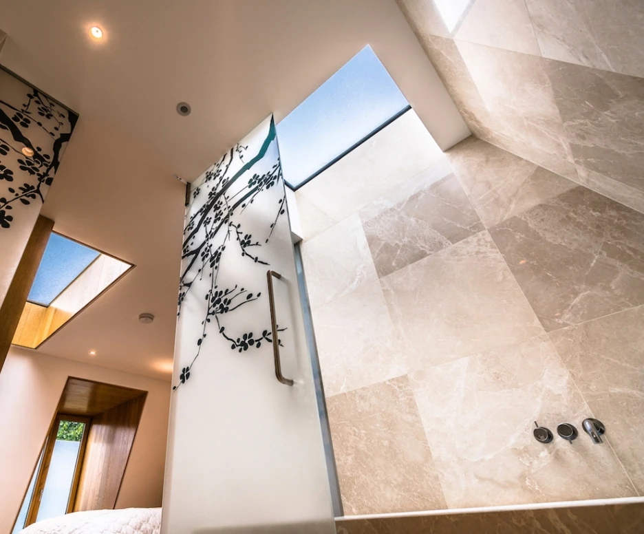 A modern bathroom with a sleek design featuring large stone tiles, a glass wall with artistic branch designs, and skylights allowing natural light to enter. The view is from below, highlighting the shower area and the visual harmony of the space.